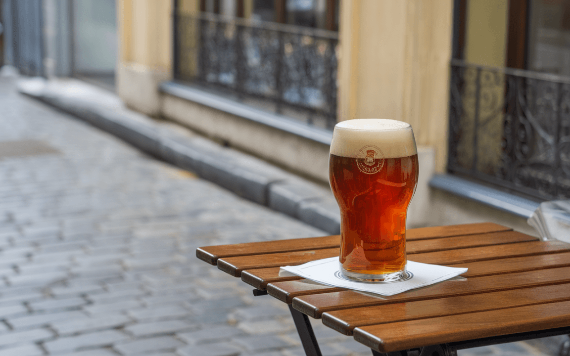 Un verre de bière ambrée sur une table en bois, placé dans une rue pavée, avec des bâtiments en arrière-plan.