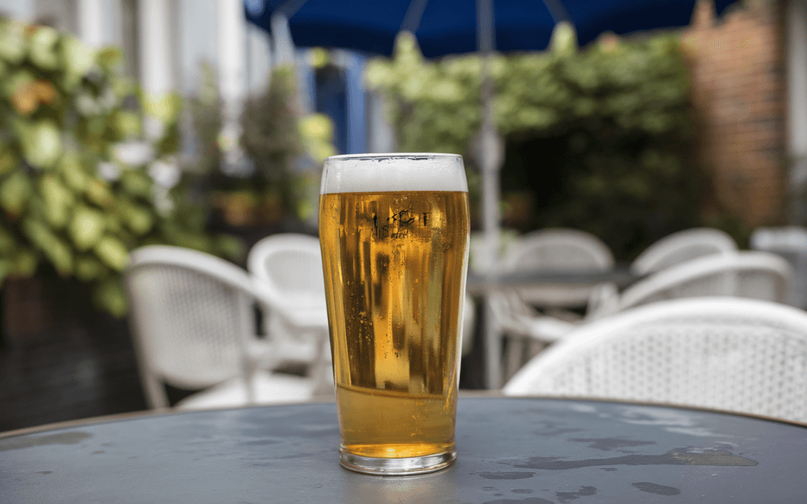 Un verre de bière froide sur une table, avec des chaises blanches en arrière-plan et un parasol bleu.