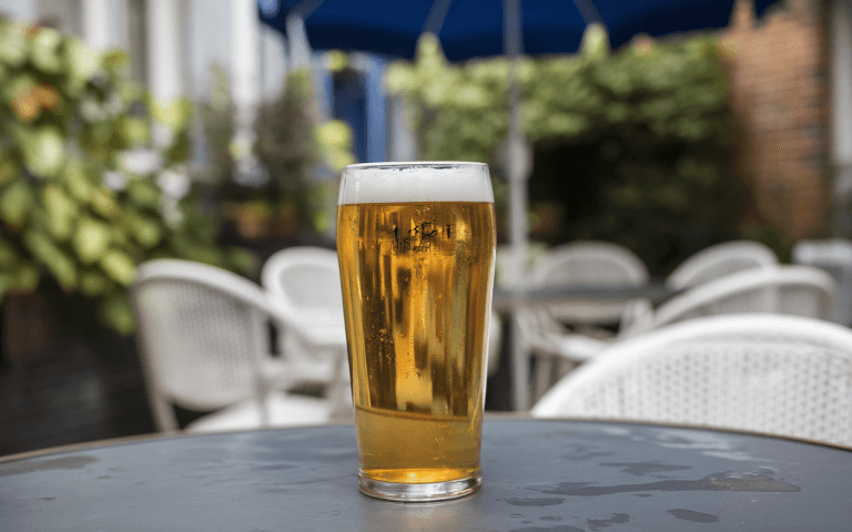 Un verre de bière froide sur une table, avec des chaises blanches en arrière-plan et un parasol bleu.