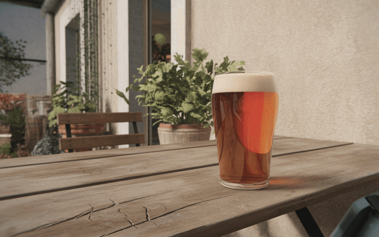 Un verre de bière brune avec mousse, posé sur une table en bois, entouré de plantes et d'une maison en arrière-plan.