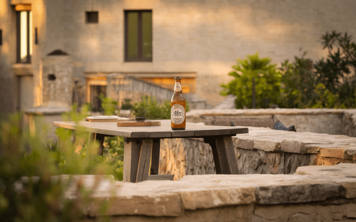 Table en extérieur avec une bière, une bouteille, un verre et des olives, vue sur des collines verdoyantes au coucher du soleil.