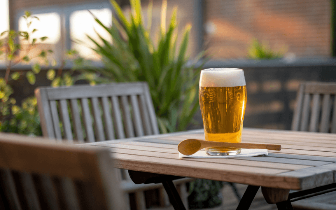 Un verre de bière dorée avec mousse est posé sur une table en bois, entouré de plantes et d’une atmosphère ensoleillée.