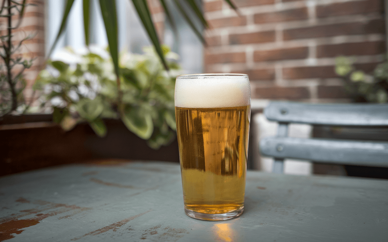 Un verre de bière blonde avec mousse, posé sur une table verte, entouré de plantes et fond de mur en briques.