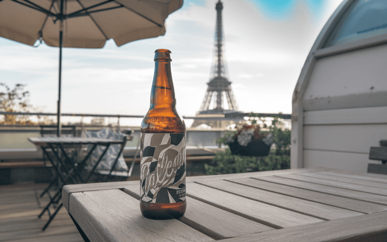 Bouteille de bière sur une table en bois, avec la tour Eiffel en arrière-plan et un ciel clair.