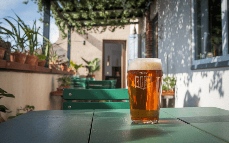 Un verre de bière artisanale sur une table verte, avec un fond de plantes et d'une maison lumineuse ensoleillée.