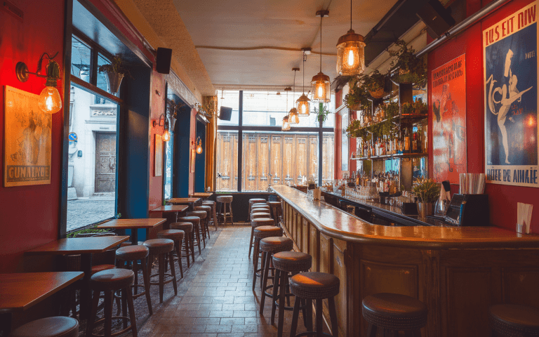 Intérieur d'un bar avec un comptoir en bois, des chaises hautes, des lumières suspendues et des affiches artistiques aux murs.