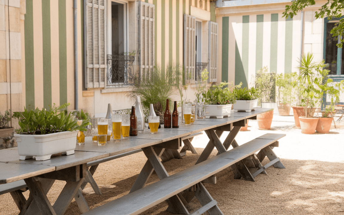 Table en bois avec des verres et bouteilles de bière, entourée de plantes, devant une façade à rayures vertes.