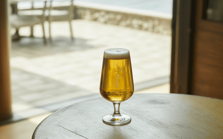 Un verre de bière dorée et mousseuse posé sur une table en bois, avec un fond flou d'extérieur.