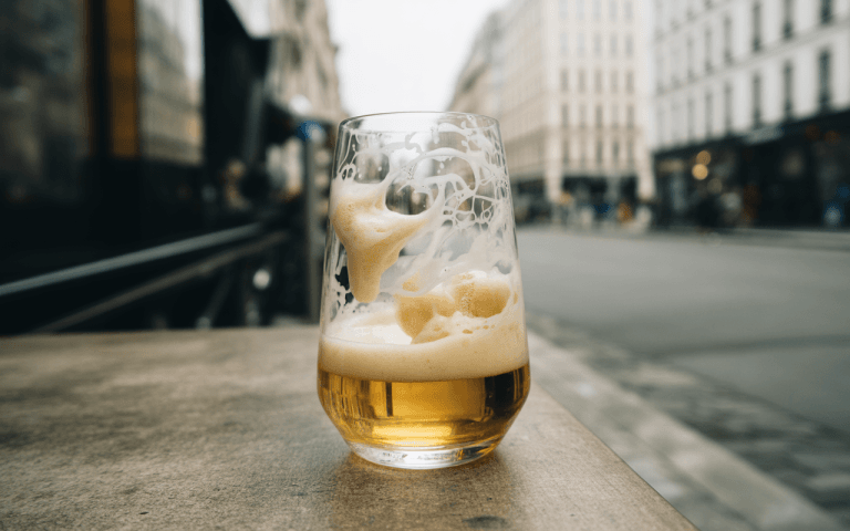 Un verre de bière avec une mousse crémeuse, posé sur une table en extérieur, avec une rue floue en arrière-plan.