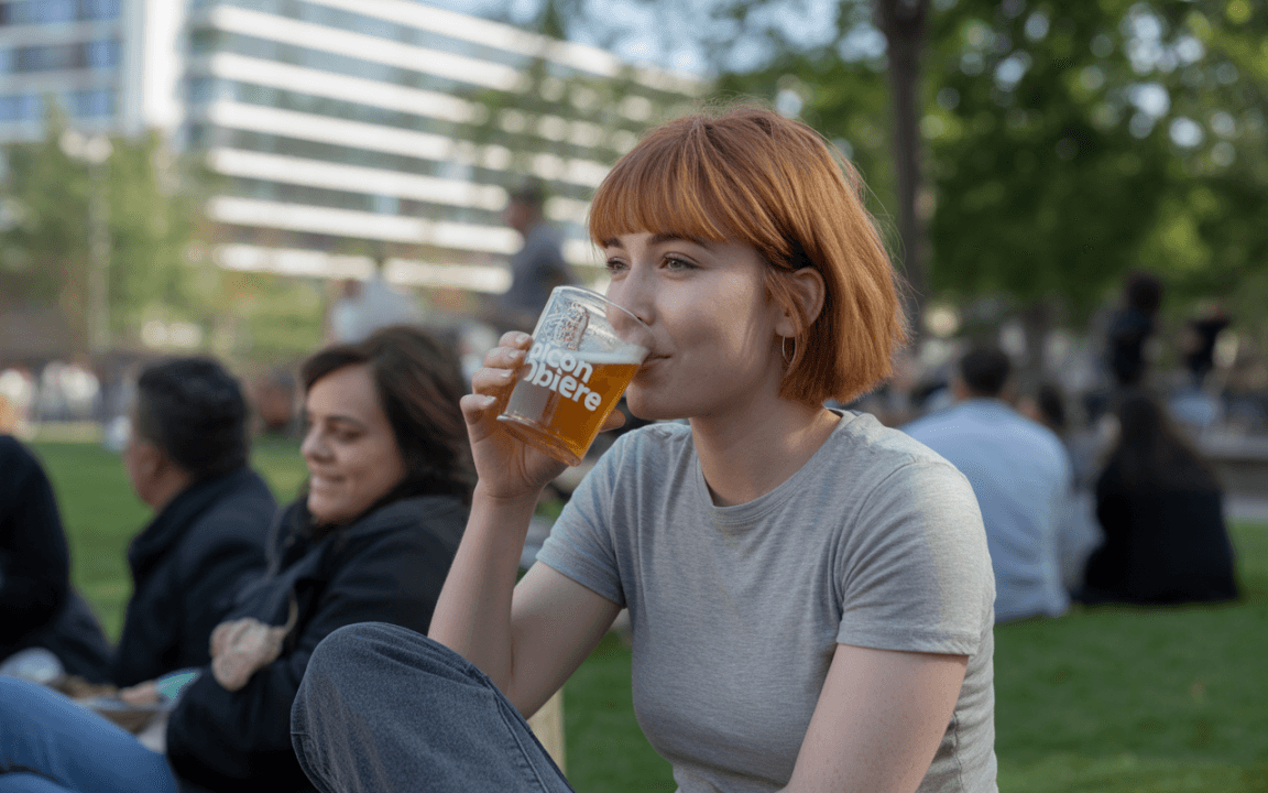 Une jeune femme aux cheveux roux boit une bière dans un parc, assise près d'autres personnes en arrière-plan.