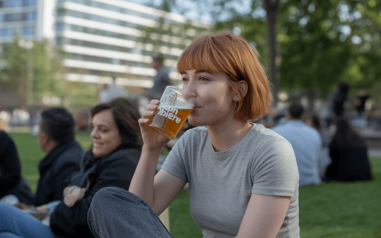 Une jeune femme aux cheveux roux boit une bière dans un parc, assise près d'autres personnes en arrière-plan.
