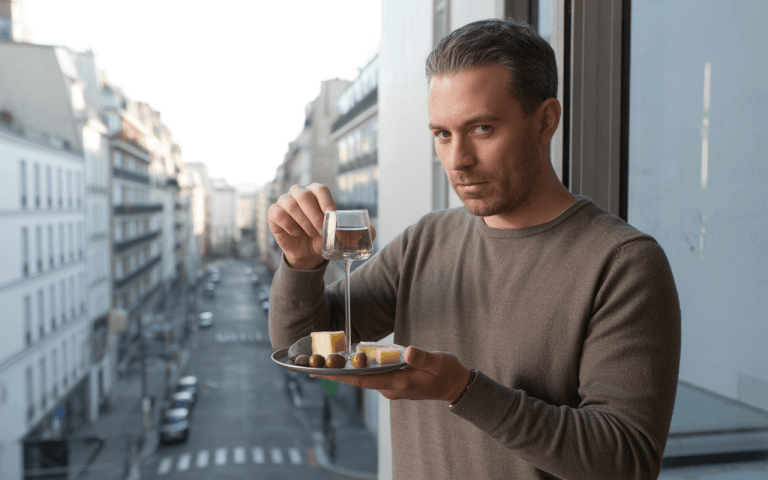 Un homme tient un plateau avec du fromage et des olives, versant une boisson depuis un verre, devant une vue urbaine.