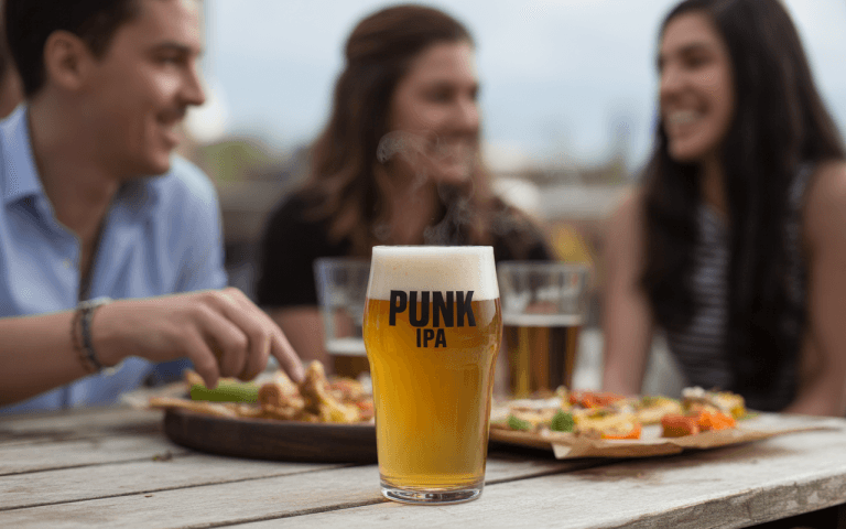 Un verre de bière IPA "PUNK" devant des amis profitant d’un repas sur une table en bois en extérieur.