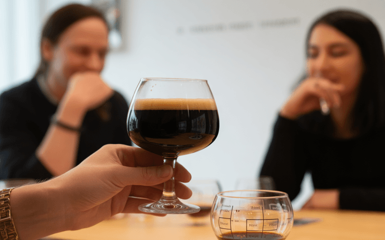 Un verre de bière noire est levé, tandis que deux personnes discutent en arrière-plan, observant la scène.