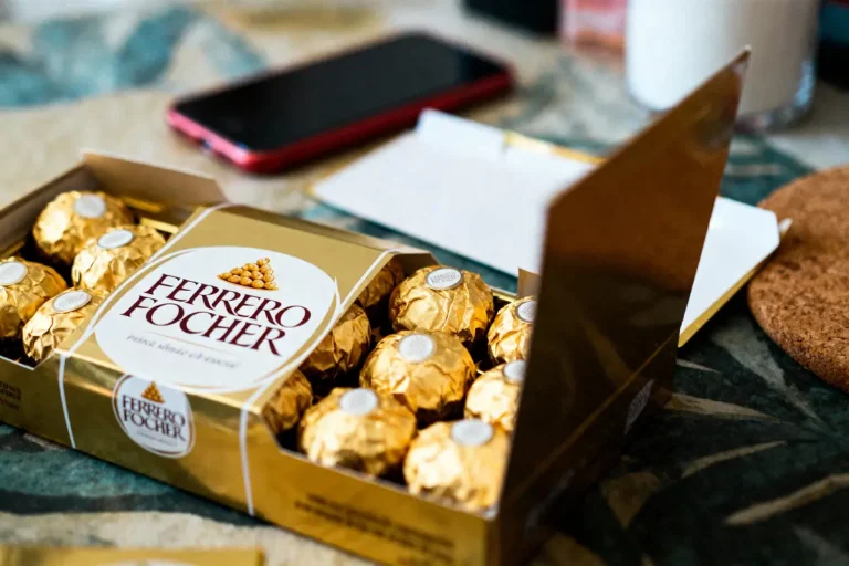 Boîte ouverte de chocolats Ferrero Rocher sur table, avec téléphone portable en arrière-plan.
