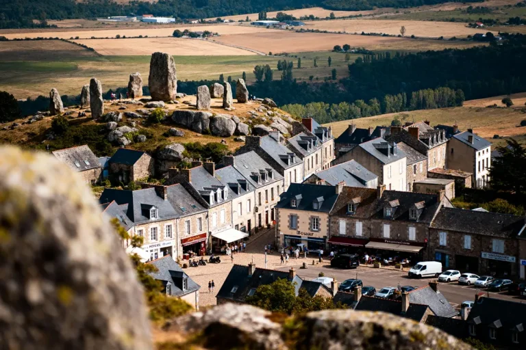 Vue de village médiéval avec maisons, dolmens et paysages vallonnés en arrière-plan.