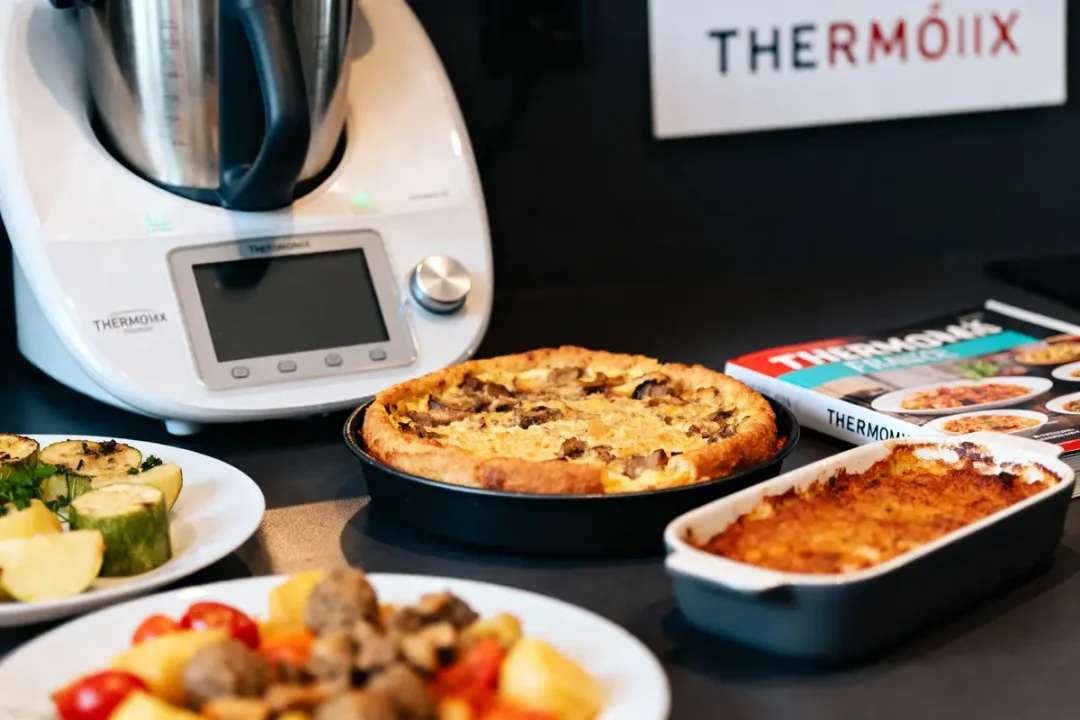 Thermomix avec plats cuisinés, livre de recettes, quiche et gratin sur plan de travail.
