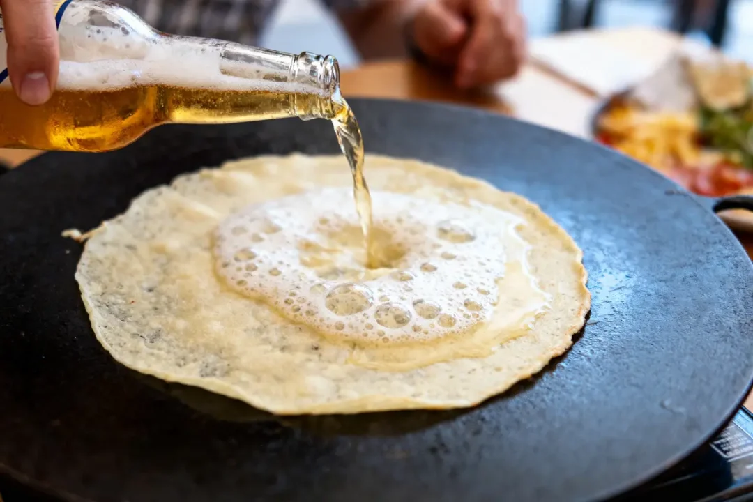 Crêpe sur poêle, bière versée dessus créant de la mousse. Main visible tenant une bouteille.