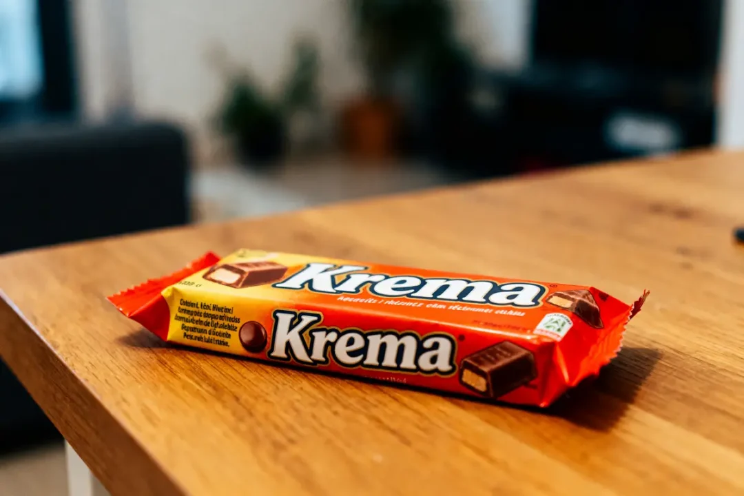 Barre chocolatée sur table en bois, emballée, marquée "Krema", arrière-plan flou avec des plantes d'intérieur.