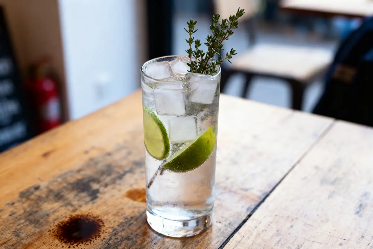 Verre de cocktail avec glaçons, rondelles de citron vert et branche de thym sur table en bois.