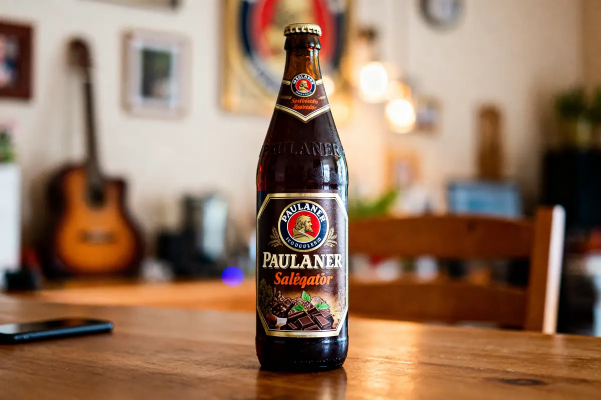 Bouteille de bière Paulaner Salégator sur table en bois, arrière-plan flou avec guitare et lumière.