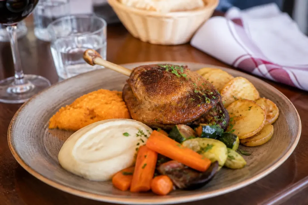 Un plat cuisse de canard rôti, carottes, purée, légumes grillés, pommes de terre. Verre de vin, serviette.