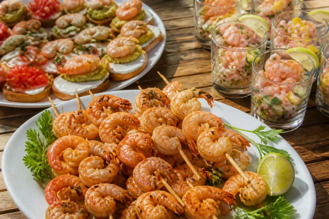 Brochettes de crevettes grillées, canapés variés et verrines de ceviche sur une table en bois.