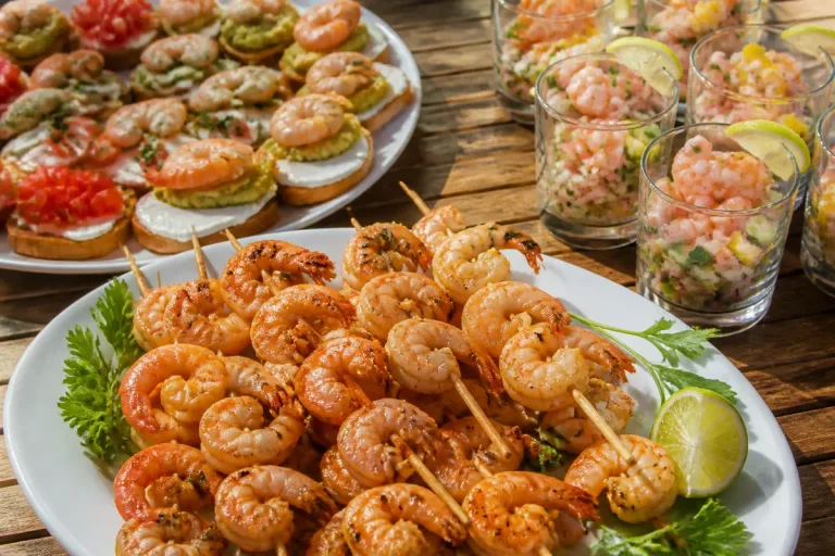Brochettes de crevettes grillées, canapés variés et verrines de ceviche sur une table en bois.