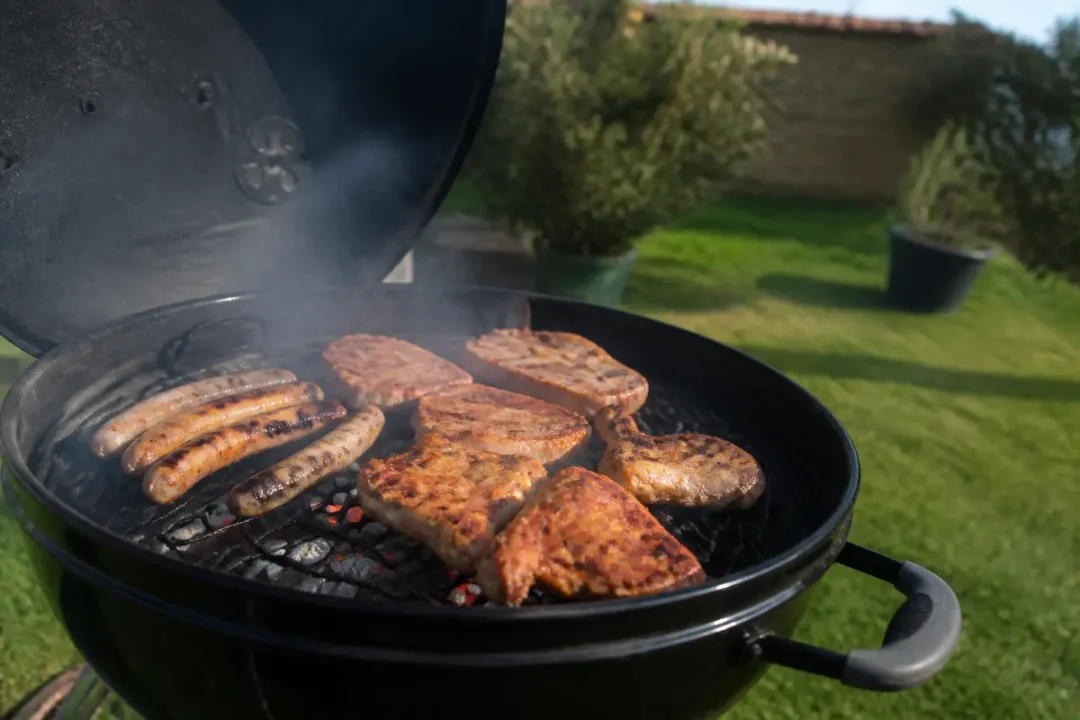 Barbecue dans jardin avec saucisses et morceaux de viande grillés sur charbon de bois, couvercle ouvert.