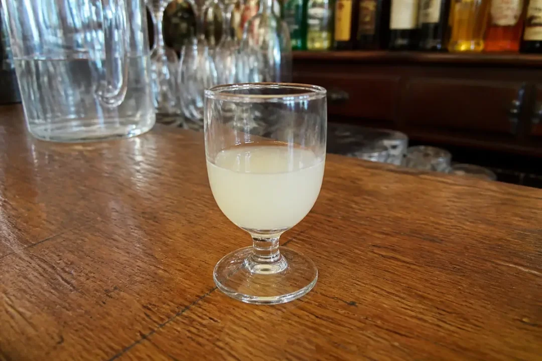 Verre de pastis posé sur un comptoir en bois, avec un fond de bouteilles et de verres.