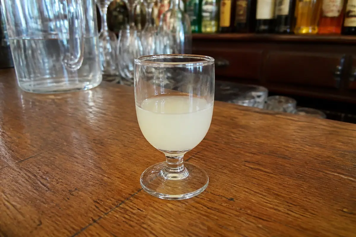 Verre de pastis posé sur un comptoir en bois, avec un fond de bouteilles et de verres.