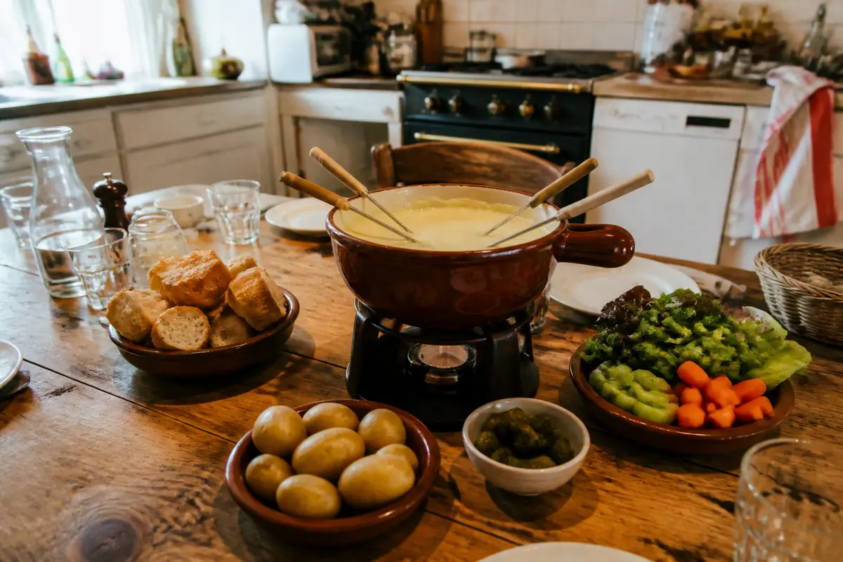 Table en bois avec fondue, légumes, pommes de terre, pain dans une cuisine rustique. Ambiance conviviale.