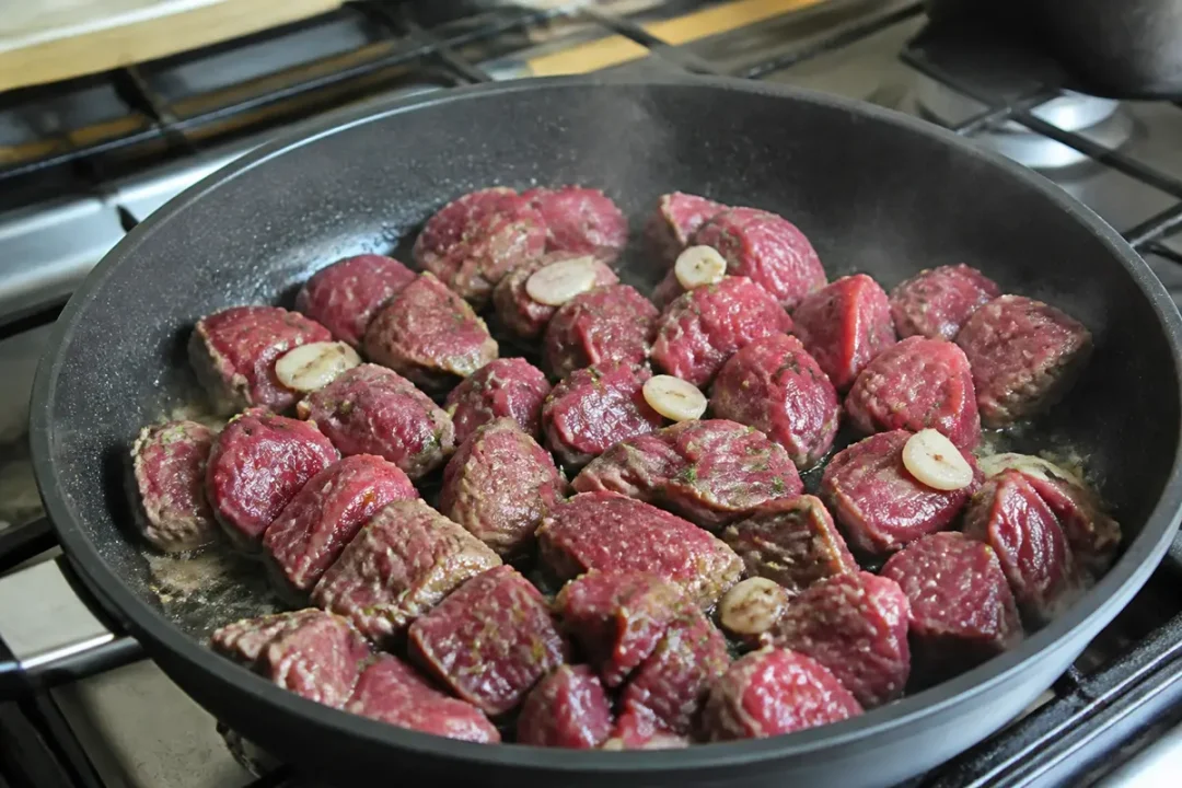 Morceaux de viande rouge cuisant dans une poêle noire, avec des tranches d'ail visible sur une cuisinière.
