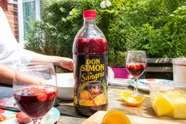 Bouteille de sangria Don Simon sur table extérieure avec verres, fruits et main d'une personne.