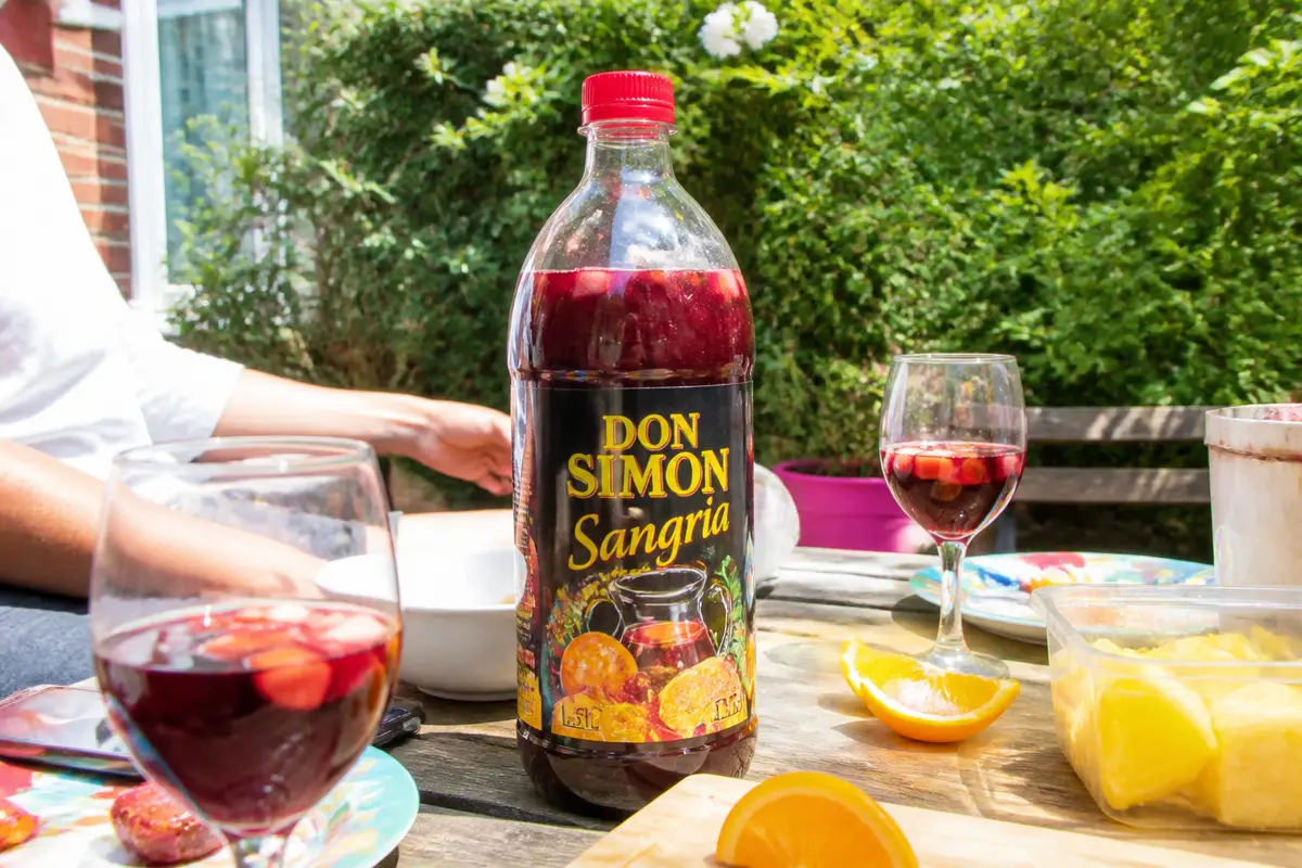 Bouteille de sangria Don Simon sur table extérieure avec verres, fruits et main d'une personne.