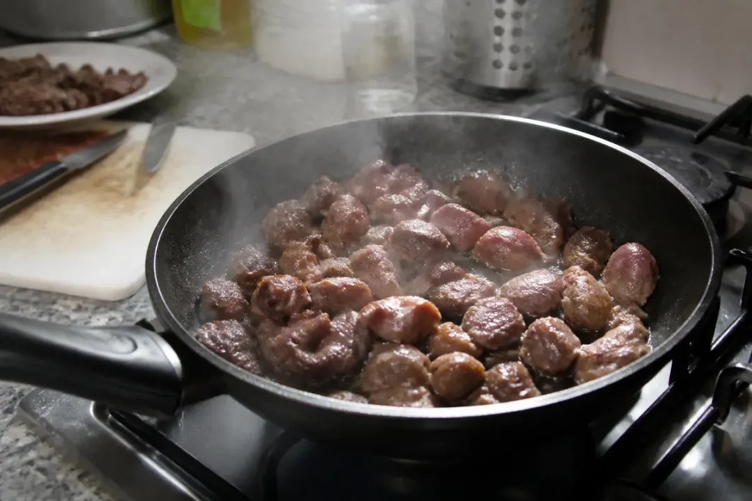 Boulettes de viande mijotant dans une poêle noire sur une cuisinière, vapeur s'élevant. Fond cuisine floue.