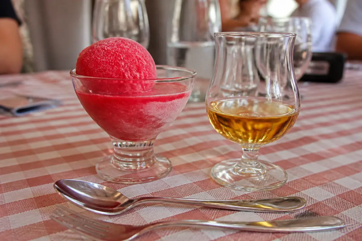 Boule de glace rouge dans une coupe à côté d'un verre de whisky sur nappe à carreaux.